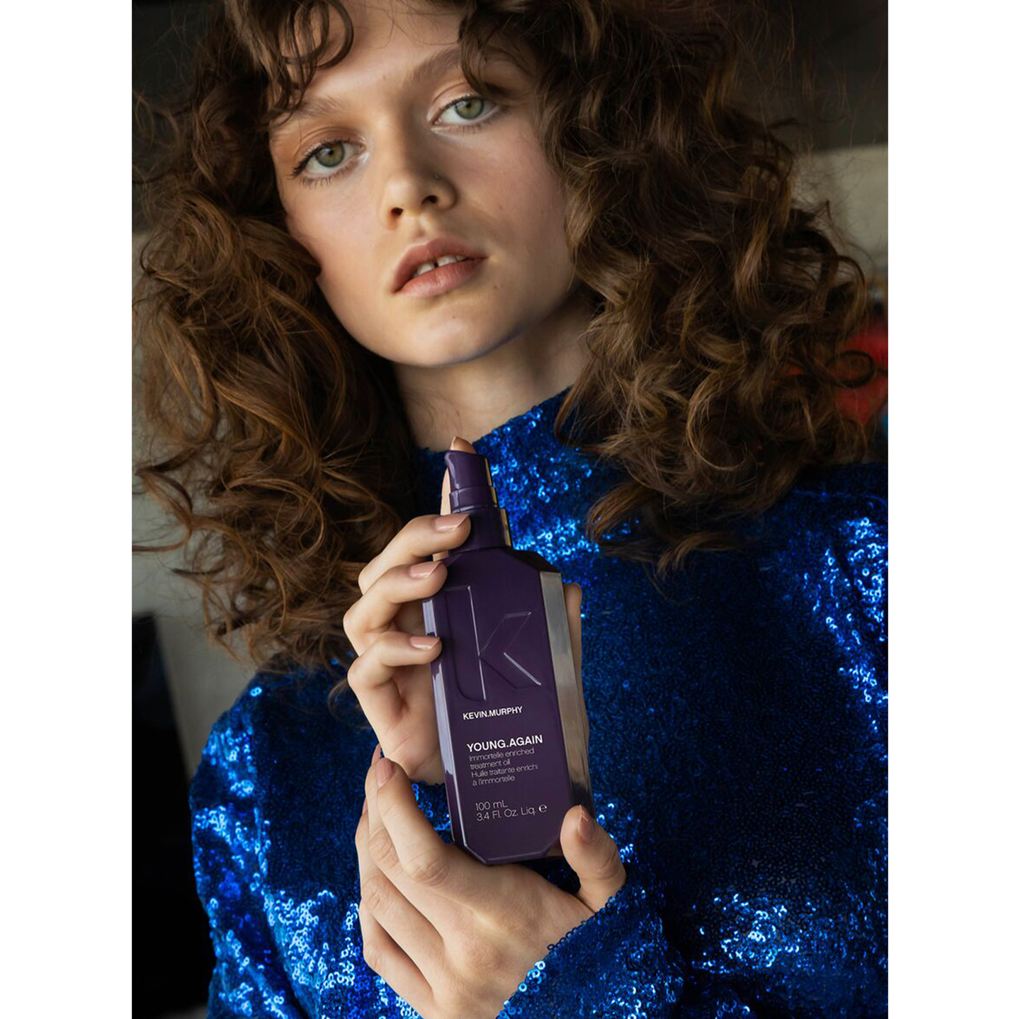 Woman with curly hair holding YOUNG.AGAIN treatment oil in blue sequin dress